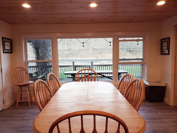Enjoy meals in this spacious dining area with a long table and six chairs, surrounded by large windows offering a view of the outdoor deck.