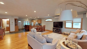 A spacious living room with a large sofa, a dining area with a wooden table and chairs, a kitchen with wooden cabinets, and a TV.