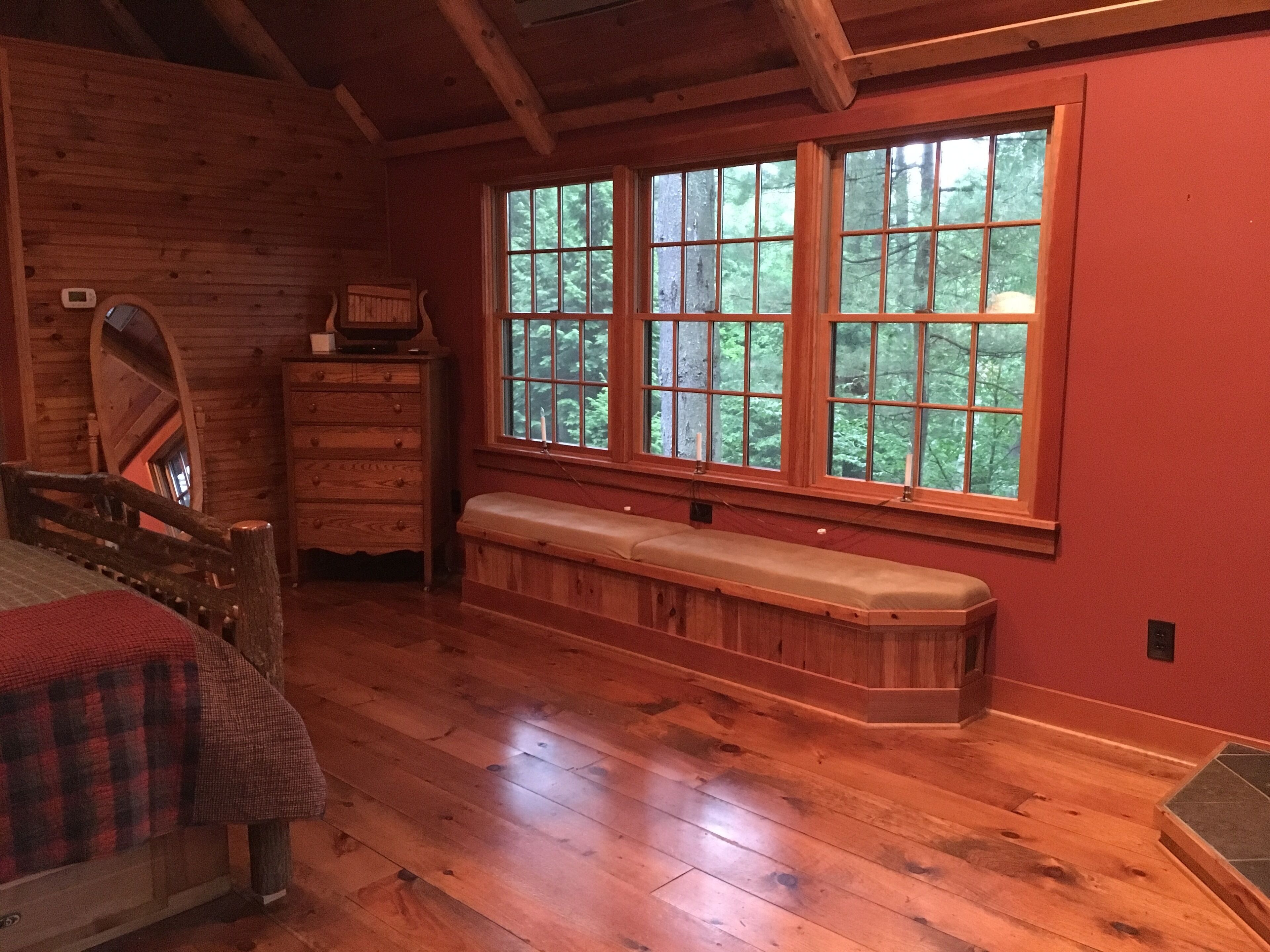 A wooden cabin room with a large window, a wooden bench, a dresser, and a bed.