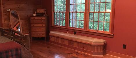 A wooden cabin room with a large window, a wooden bench, a dresser, and a bed.