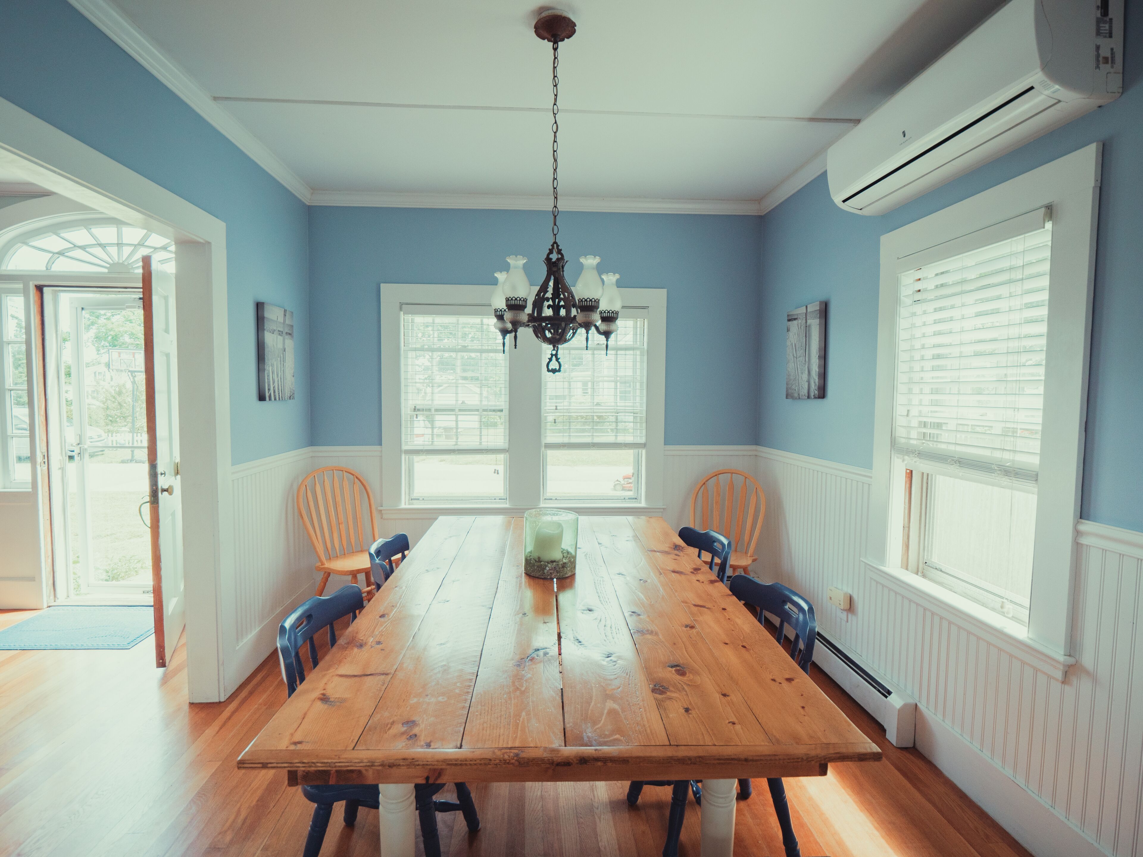 A dining room with a wooden table, two chairs, a chandelier, and large windows.