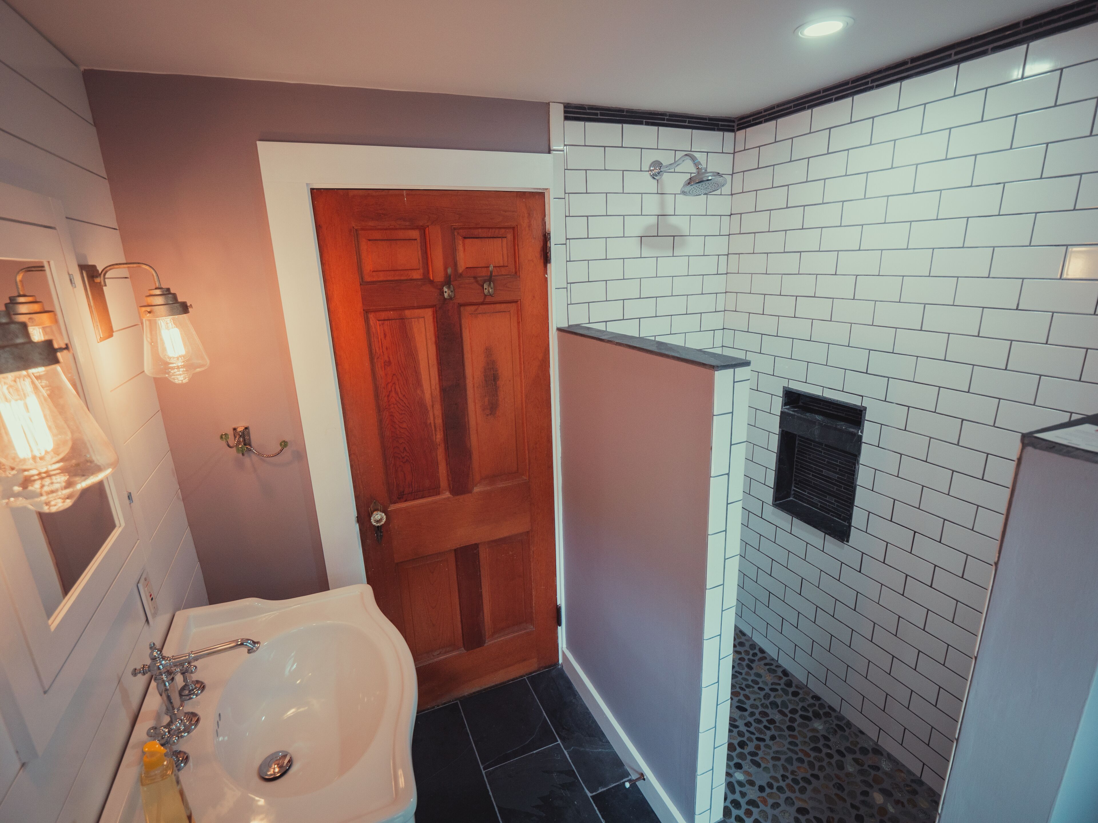 Une salle de bain avec une baignoire blanche, une porte en bois et un espace douche avec des carreaux blancs et une grille d’aération noire.