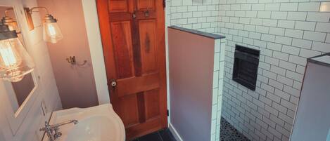 Une salle de bain avec une baignoire blanche, une porte en bois et un espace douche avec des carreaux blancs et une grille d’aération noire.