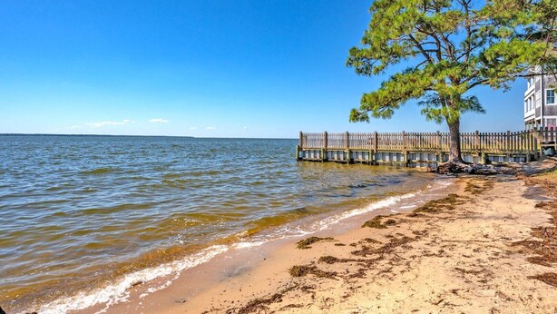 En strand med en träbrygga, ett träd och en byggnad vid vattnet.