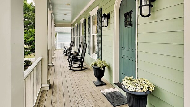 A covered porch with wooden railings, rocking chairs, a potted plant, and a green door with a window.