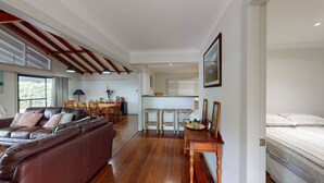 Amplia sala de estar con un sofá de cuero marrón, una mesa de comedor de madera y sillas. Hay una zona de cocina con ventana y una cama en la habitación contigua.