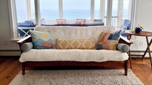 Una sala de estar acogedora con un sofá blanco mullido, un tapete estampado y vistas al océano a través de amplias ventanas.