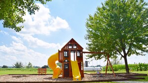 Une aire de jeux avec un toboggan jaune et des balançoires, entourée d’arbres et d’un champ d’herbe.