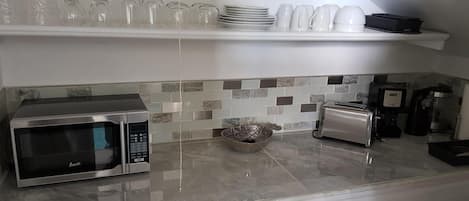 A modern kitchen with a microwave, toaster, and coffee maker. There are shelves with plates and glasses above the counter.
