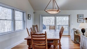 Comedor con una amplia mesa de madera y sillas, ventanas grandes y vistas a árboles.