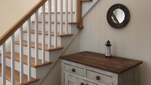 Una escalera con escalones de madera y barandillas blancas, un mueble de madera pequeño con cajones, un espejo redondo y un cuadro enmarcado en la pared.