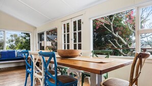 Un coin repas avec une table en bois, des chaises bleues et blanches, et une vue sur les arbres à travers de grandes fenêtres.