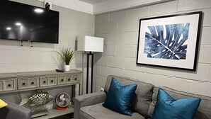 A modern living room with a grey sofa, blue cushions, a mounted TV, a white cabinet, a floor lamp, and a framed abstract art piece. - SEAS THE DAY: Charming Folly Beach Apartment awaits your arrival! (Folly Beach)