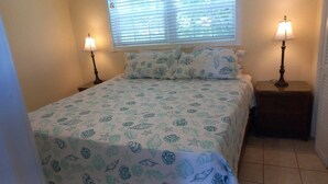 Un dormitorio con cama, dos mesitas de noche con lámparas, una ventana con persianas y una colcha estampada.