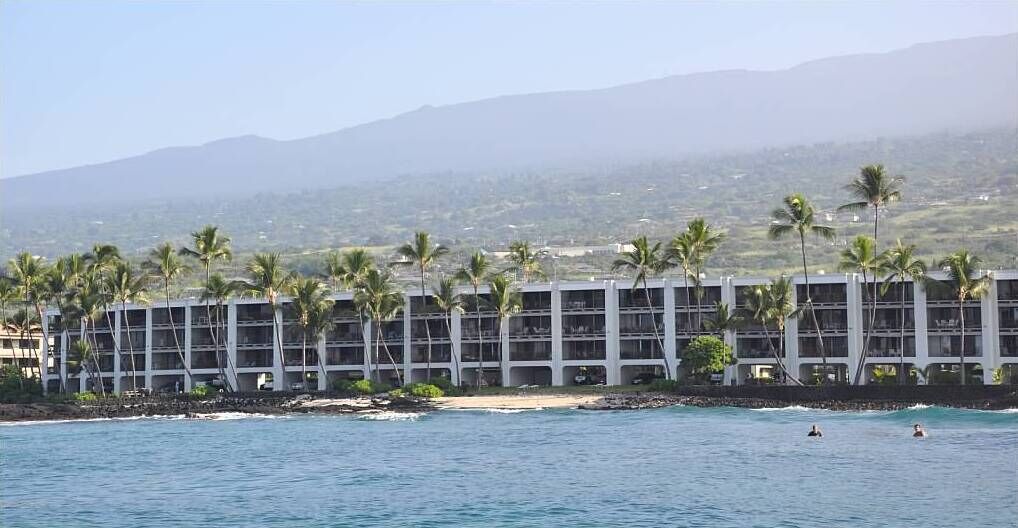 A beachfront hotel with multiple floors, palm trees, and a view of the ocean and mountains.