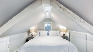 Une chambre avec un plafond en pente, un lit avec une literie blanche, deux tables de chevet avec des lampes et une fenêtre donnant sur un beau paysage.