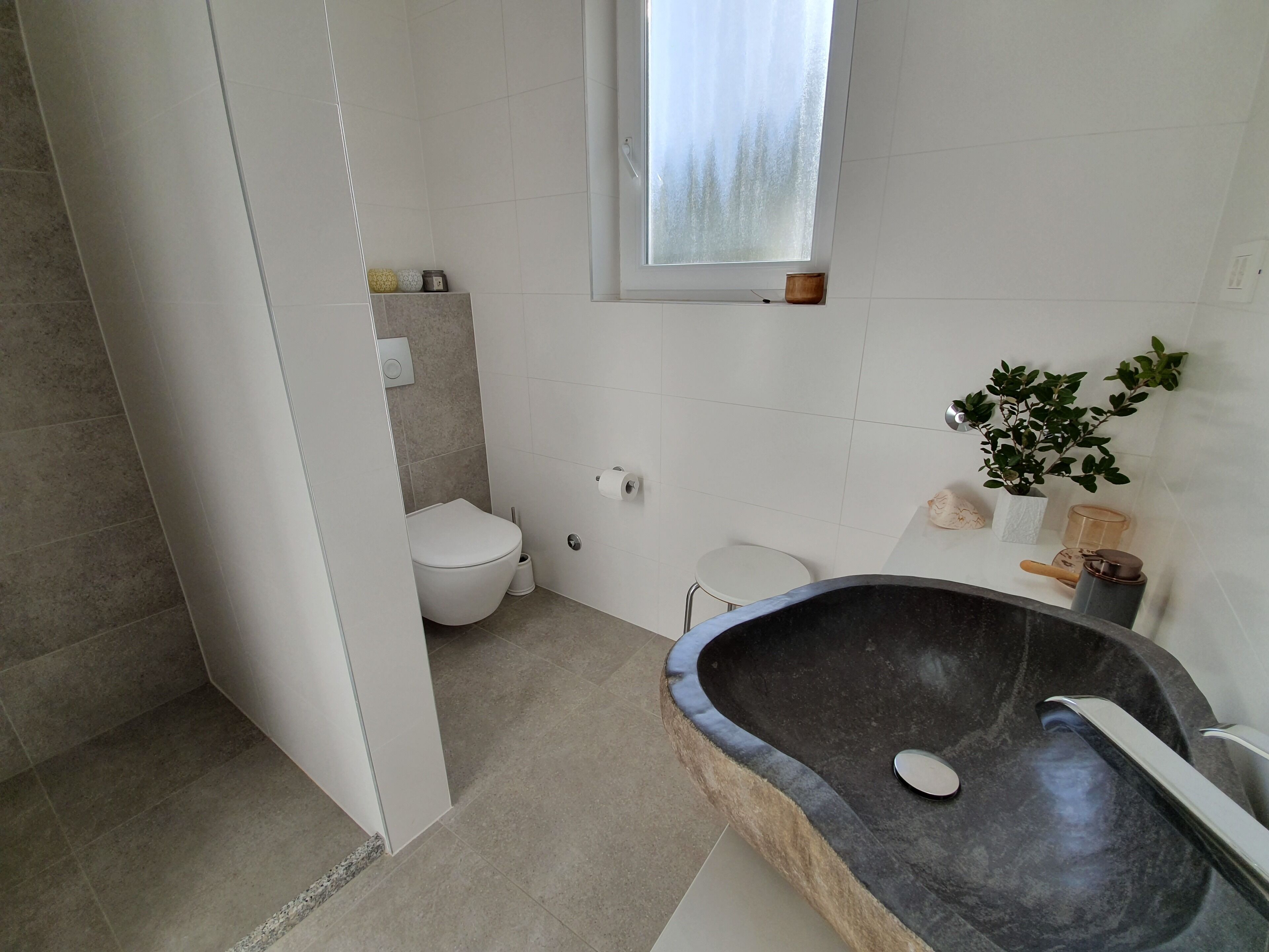A bathroom with a toilet, a sink with a basin, a small stool, and a potted plant.