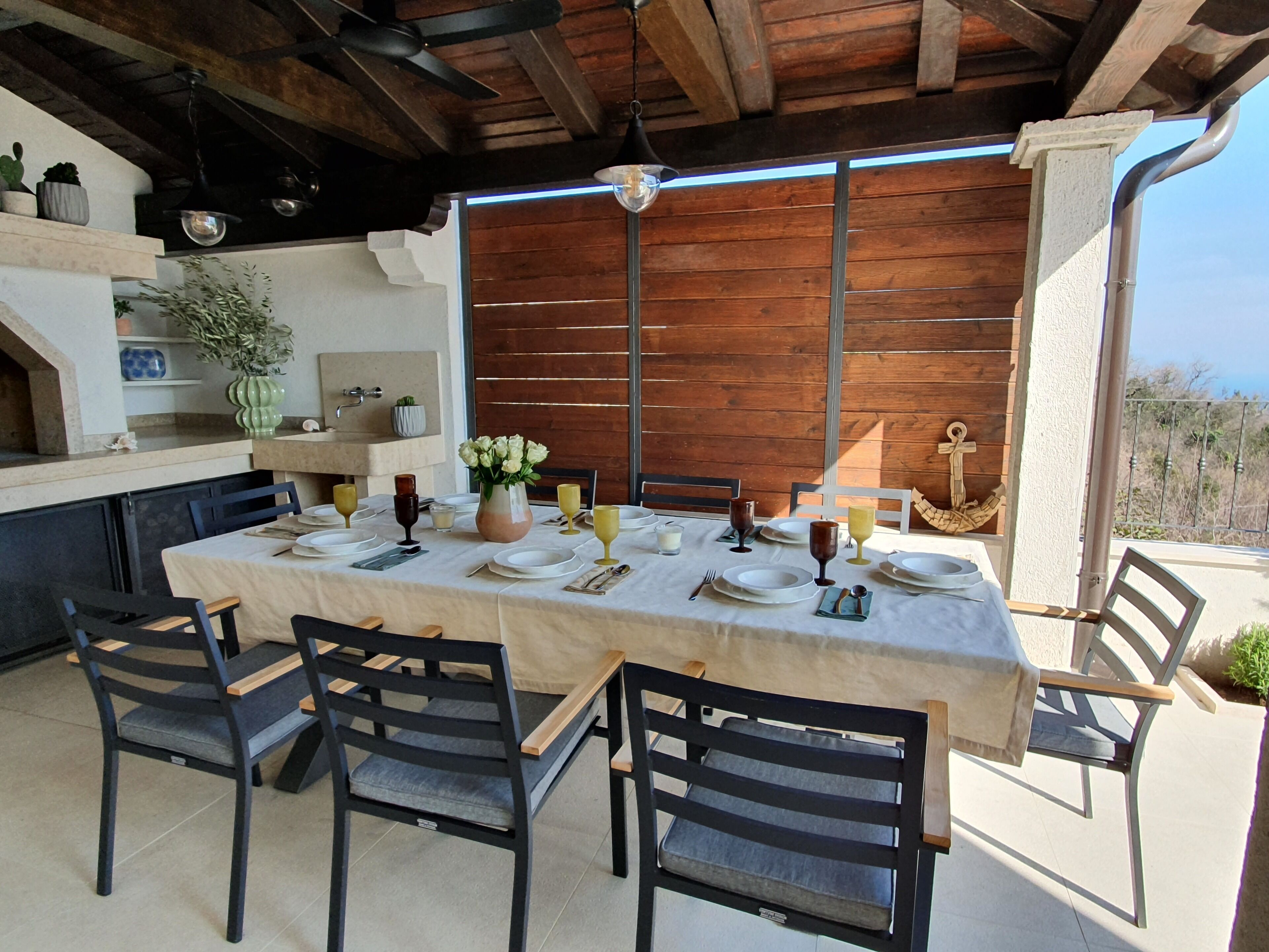 Un espacio de comedor al aire libre con una mesa dispuesta para cuatro personas, rodeada de sillas, bajo una pérgola de madera con vistas al paisaje.