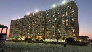 Un hotel de gran altura con varios balcones, arena en primer plano y cielo despejado.