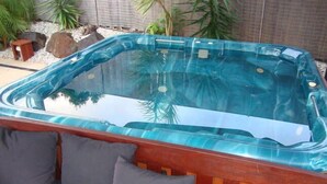 A hot tub with a glass enclosure, surrounded by a wooden deck and gray cushions. - Kooroora House -Spa Bath-Pool- Billard Table (Robina)