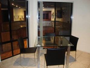 A dining area with a glass table and four chairs, a kitchen visible through a glass partition, and a modern interior design. - Kooroora House -Spa Bath-Pool- Billard Table (Robina)