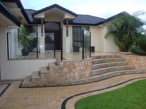 A modern house with a stone staircase leading to a glass door entrance, surrounded by a well-maintained lawn and palm trees. - Kooroora House -Spa Bath-Pool- Billard Table (Robina)