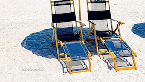 Una playa con dos sillas de playa bajo una sombrilla azul, un edificio al fondo y dunas de arena.