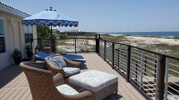 Eine Holzterrasse mit Korbmöbeln, einem blauen Sonnenschirm und Blick auf den Strand.