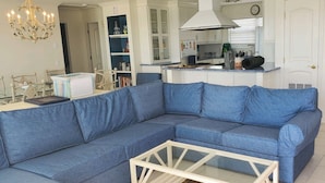 Ein Wohnzimmer mit blauem Ecksofa, einem Glastisch und einer Küchenzeile mit weißen Schränken und einem Herd.