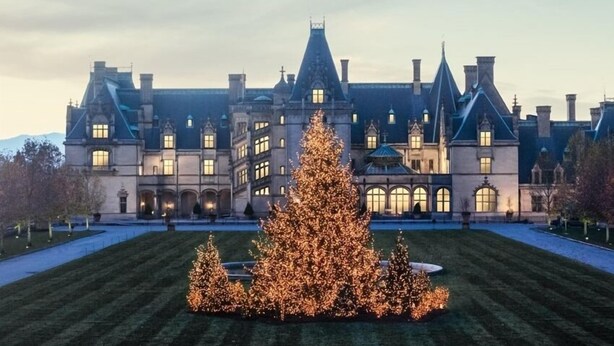 A large, historic building with multiple chimneys and arched windows, surrounded by a well-manicured lawn and illuminated Christmas trees.