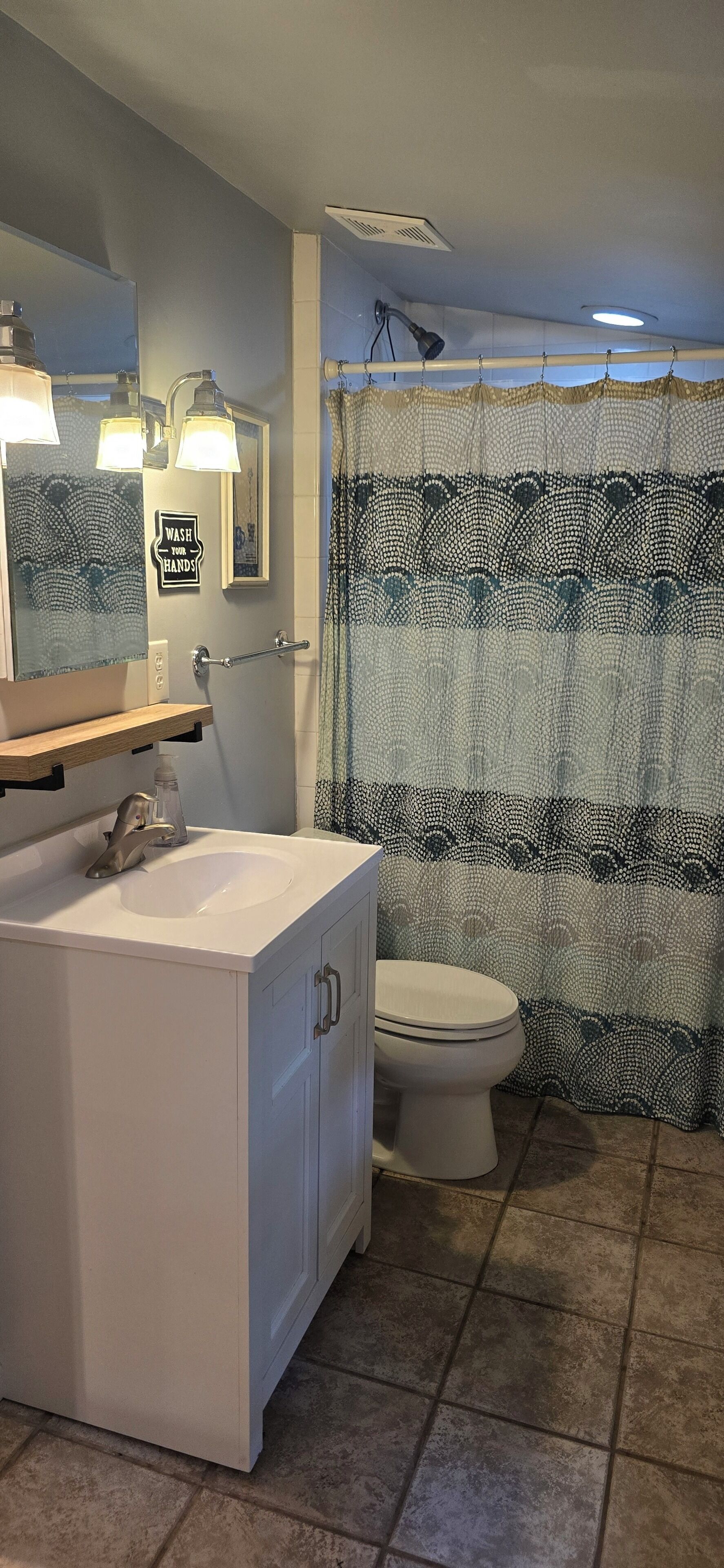 Un baño con lavamanos blanco, inodoro, cortina de ducha con diseño de olas y espejo con luces.