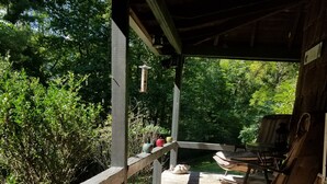 A wooden deck with a railing, surrounded by greenery, and a chair with a cushion.