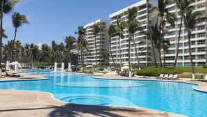 Uma piscina grande com espreguiçadeiras e palmeiras, rodeada por um edifício de hotel com vários andares.