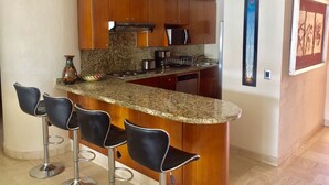 Cozinha com armários de madeira, bancada de granito, geladeira de aço inoxidável e área de bar com quatro banquetas.