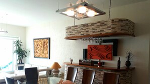 Uma área de refeições com mesa de vidro, cadeiras, uma tigela decorativa, luminária na parede e parede de tijolos.