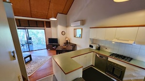 A compact kitchen with a central island, a stove, and a microwave. There is a dining area with a table and chairs, and a television in the background. - Sapphire Villa 535, Kingfisher Bay Resort, K'gari (Fraser Island)