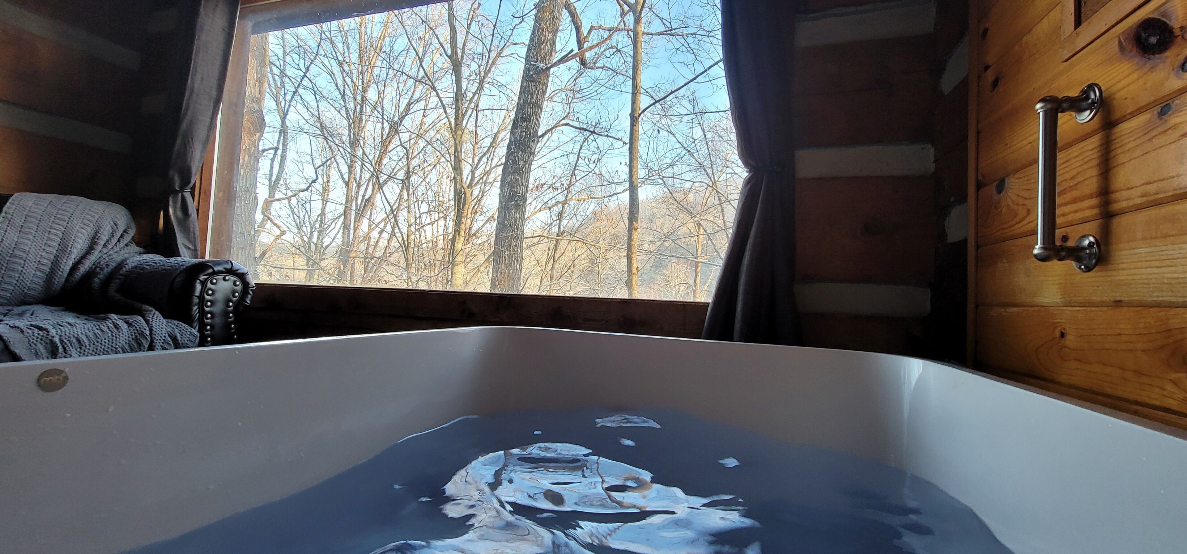 A cabin interior with a bathtub, wooden walls, and a window with a view of leafless trees.
