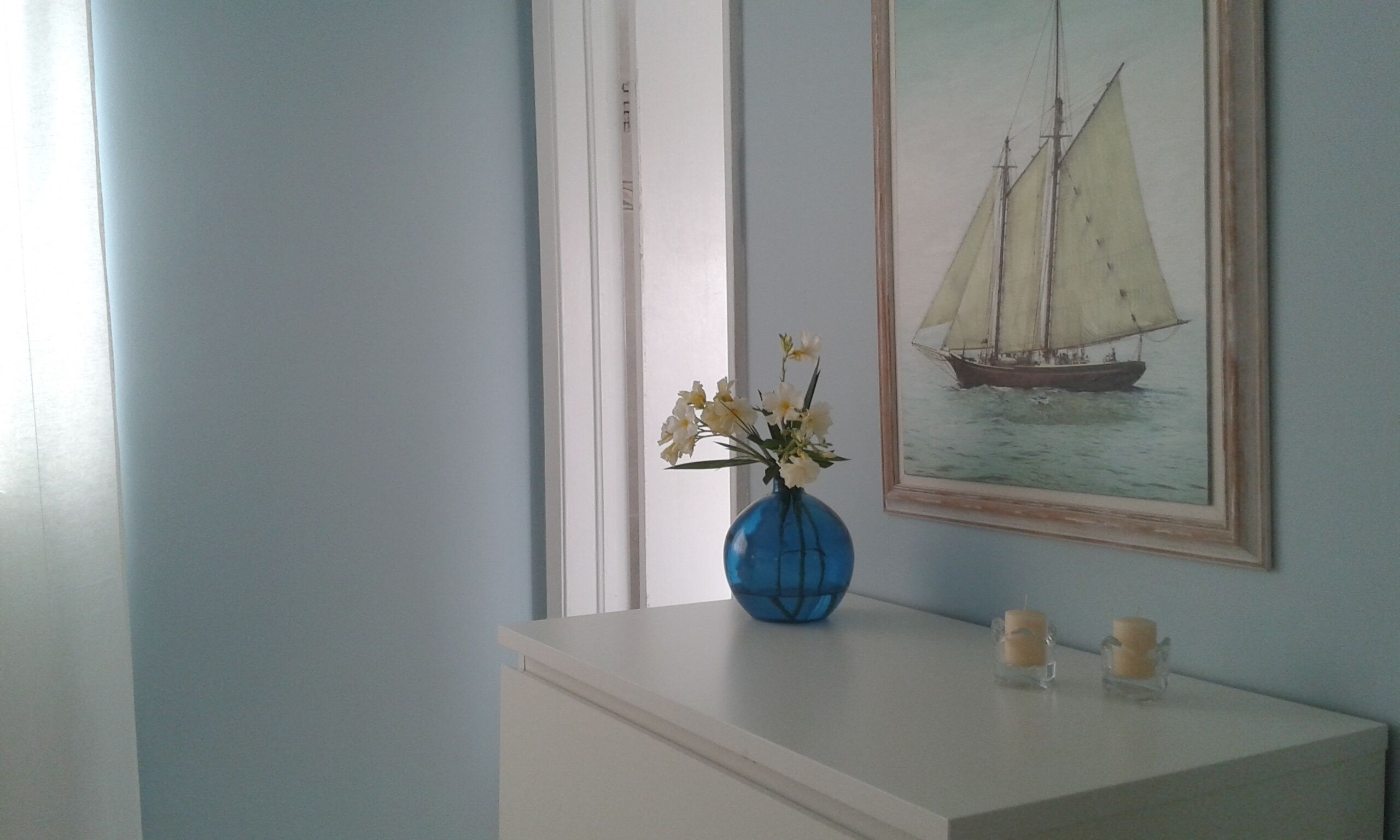 Ein Zimmer mit einem weißen Schminktisch, einer blauen Vase mit Blumen, zwei Kerzen und einem gerahmten Bild eines Segelbootes an der Wand.