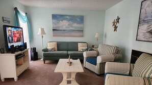 Un salon avec un canapé vert, des fauteuils à rayures, une table basse et un téléviseur posé sur un meuble.