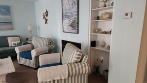 Un salon avec des fauteuils à rayures, un canapé vert, une cheminée et une bibliothèque agrémentée d’objets décoratifs.