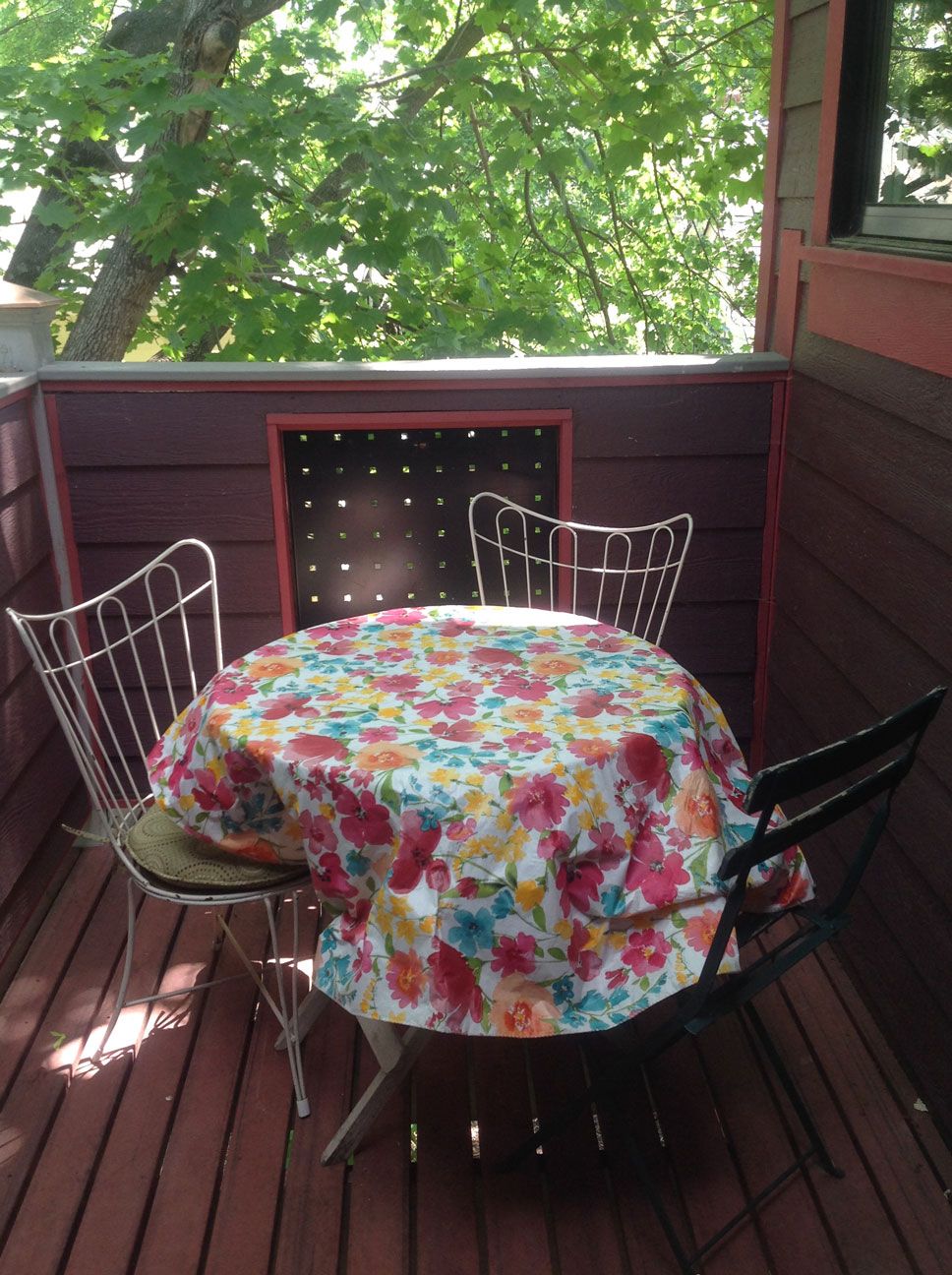 Une table ronde recouverte d’une nappe à motifs floraux, entourée de quatre chaises en métal, sur une terrasse en bois offrant une vue sur les arbres.