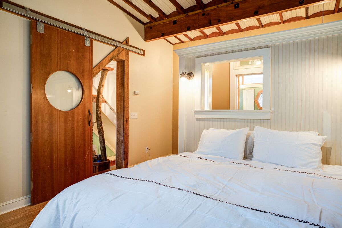 Une chambre avec un grand lit, une porte en bois ornée d’un miroir rond, un escalier en bois et une fenêtre avec des stores.