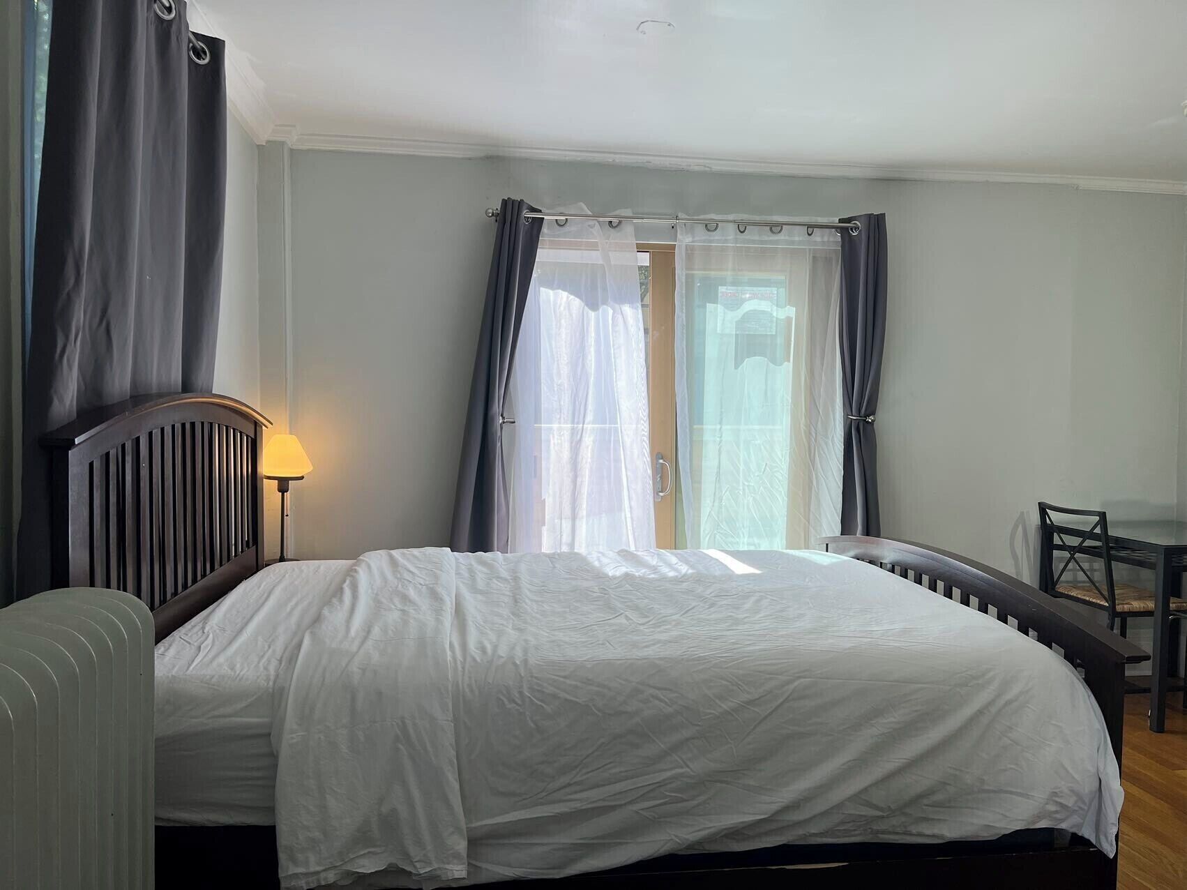 A bedroom with a bed, a bedside table, a lamp, a chair, and a view of the outdoors through a window with curtains.