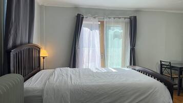 A bedroom with a bed, a bedside table, a lamp, a chair, and a view of the outdoors through a window with curtains.