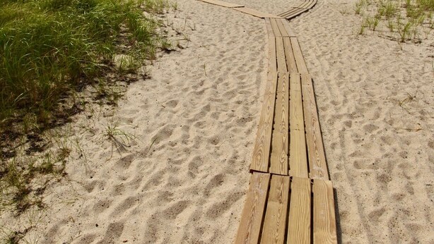 Una passerella in legno che conduce a una spiaggia con sabbia ed erba ai lati.