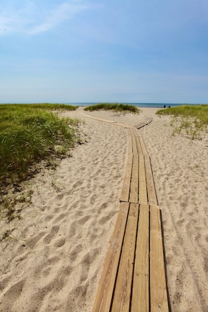 Ein hölzerner Steg führt zu einem Strand mit Sand und Gras an beiden Seiten.