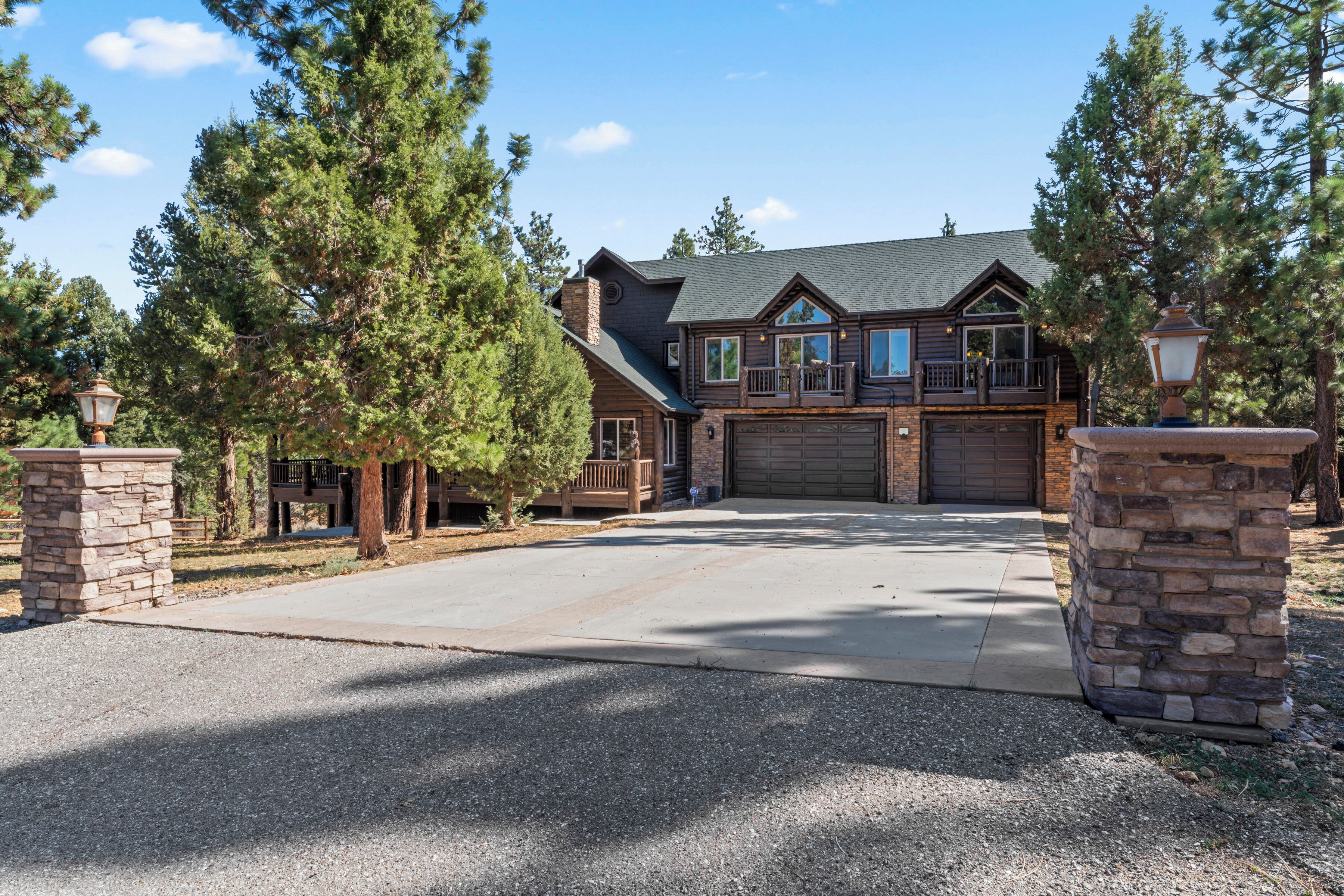 A two-story log cabin with a garage, surrounded by tall pine trees.