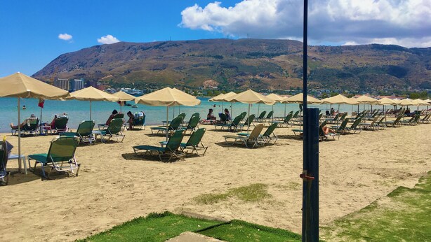 Une plage bordée de rangées de chaises longues et de parasols, un ciel dégagé et un paysage vallonné en arrière-plan.
