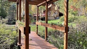 Una terraza de madera con una estructura de pérgola, rodeada de abundante vegetación y una cabaña de madera al fondo.