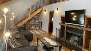Amplia sala de estar con techo alto, escalera, chimenea, televisor grande y estantería.